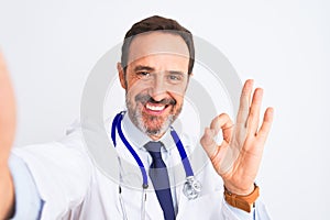 Middle age doctor man wearing stethoscope make selfie over isolated white background doing ok sign with fingers, excellent symbol