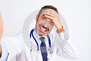 Middle age doctor man wearing stethoscope make selfie over isolated white background stressed with hand on head, shocked with
