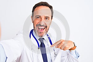 Middle age doctor man wearing stethoscope make selfie over isolated white background with surprise face pointing finger to himself