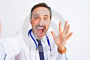 Middle age doctor man wearing stethoscope make selfie over isolated white background very happy and excited, winner expression