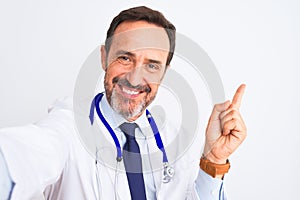 Middle age doctor man wearing stethoscope make selfie over isolated white background very happy pointing with hand and finger to