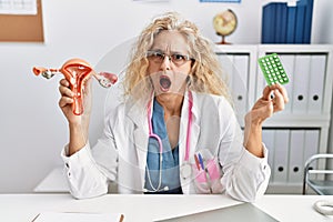 Middle age doctor woman holding birth control pills in shock face, looking skeptical and sarcastic, surprised with open mouth