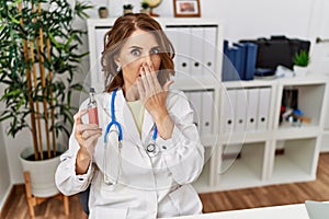 Middle age doctor woman holding electronic cigarette at medical clinic covering mouth with hand, shocked and afraid for mistake