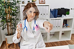 Middle age doctor woman holding electronic cigarette at medical clinic pointing with finger to the camera and to you, confident