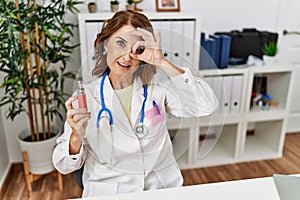 Middle age doctor woman holding electronic cigarette at medical clinic smiling happy doing ok sign with hand on eye looking