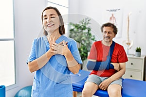 Middle age doctor woman with patient with arm injury at rehabilitation clinic smiling with hands on chest, eyes closed with