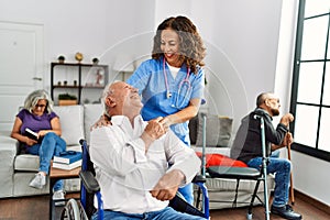 Middle age doctor woman supporting retired man sitting on wheelchair at nursing home