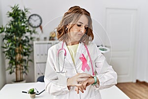 Middle age doctor woman wearing pink cancer ribbon on uniform checking the time on wrist watch, relaxed and confident