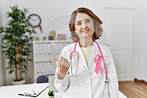 Middle age doctor woman wearing pink cancer ribbon on uniform pointing to the back behind with hand and thumbs up, smiling