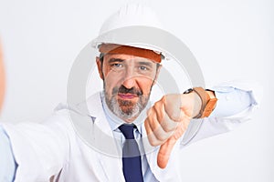 Middle age engineer man wearing helmet make selfie over isolated white background with angry face, negative sign showing dislike