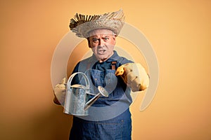 Middle age farmer man wearing apron and hat holding watering can over yellow background annoyed and frustrated shouting with