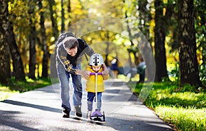 Middle age father showing his toddler son how to ride a scooter in a autumn park