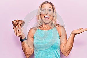 Middle age fit blonde woman holding bowl with almonds celebrating achievement with happy smile and winner expression with raised