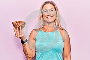 Middle age fit blonde woman holding bowl with almonds looking positive and happy standing and smiling with a confident smile