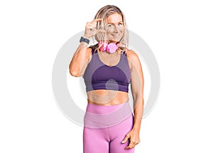 Middle age fit blonde woman wearing gym clothes and using headphones smiling and confident gesturing with hand doing small size