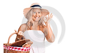 Middle age fit blonde woman wearing summer hat and holding picnic wicker basket with bread doing ok sign with fingers, smiling