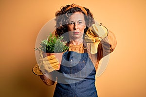 Middle age gardener woman wearing apron holding plant pot over isolated yellow background with angry face, negative sign showing