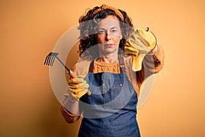 Middle age gardener woman wearing apron holding rake over isolated yellow background with angry face, negative sign showing