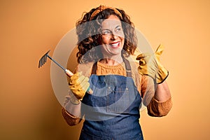 Middle age gardener woman wearing apron holding rake over isolated yellow background pointing and showing with thumb up to the