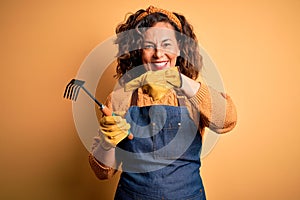 Middle age gardener woman wearing apron holding rake over isolated yellow background very happy pointing with hand and finger