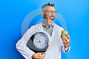 Middle age grey-haired man as nutritionist doctor holding weighing machine and green apple angry and mad screaming frustrated and