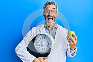 Middle age grey-haired man as nutritionist doctor holding weighing machine and green apple celebrating crazy and amazed for