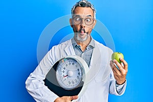 Middle age grey-haired man as nutritionist doctor holding weighing machine and green apple making fish face with mouth and