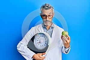 Middle age grey-haired man as nutritionist doctor holding weighing machine and green apple skeptic and nervous, frowning upset