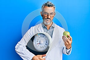 Middle age grey-haired man as nutritionist doctor holding weighing machine and green apple skeptic and nervous, frowning upset