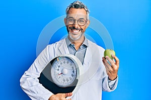 Middle age grey-haired man as nutritionist doctor holding weighing machine and green apple smiling with a happy and cool smile on