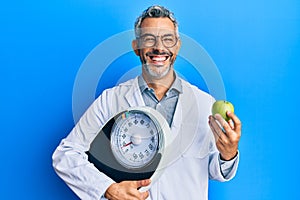 Middle age grey-haired man as nutritionist doctor holding weighing machine and green apple smiling and laughing hard out loud