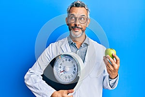 Middle age grey-haired man as nutritionist doctor holding weighing machine and green apple smiling looking to the side and staring