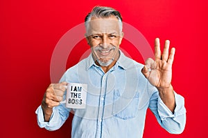 Middle age grey-haired man drinking from i am the boss coffee cup doing ok sign with fingers, smiling friendly gesturing excellent