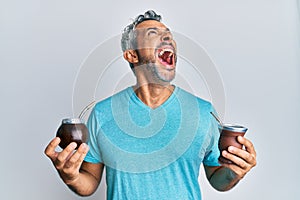 Middle age grey-haired man drinking mate infusion angry and mad screaming frustrated and furious, shouting with anger looking up