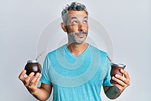 Middle age grey-haired man drinking mate infusion smiling looking to the side and staring away thinking