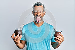 Middle age grey-haired man drinking mate infusion sticking tongue out happy with funny expression