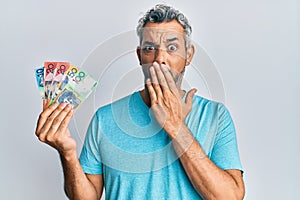 Middle age grey-haired man holding australian dollars covering mouth with hand, shocked and afraid for mistake