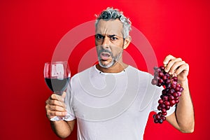 Middle age grey-haired man holding branch of fresh grapes and red wine in shock face, looking skeptical and sarcastic, surprised
