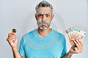 Middle age grey-haired man holding bunch of 50 euro banknotes and bitcoin puffing cheeks with funny face