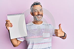 Middle age grey-haired man holding empty notebook smiling happy and positive, thumb up doing excellent and approval sign