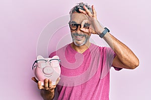 Middle age grey-haired man holding piggy bank with glasses smiling happy doing ok sign with hand on eye looking through fingers