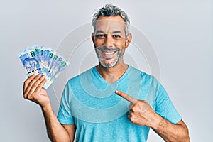 Middle age grey-haired man holding south african rands banknotes smiling happy pointing with hand and finger