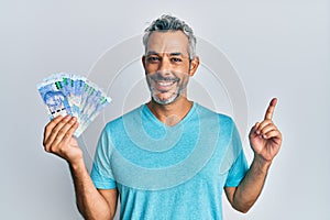Middle age grey-haired man holding south african rands banknotes smiling happy pointing with hand and finger to the side