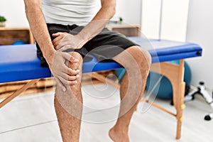 Middle age grey-haired man patient suffering for knee injury sitting on massage table at rehab clinic