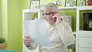 Middle age grey-haired man reading document talking on smartphone at home