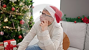 Middle age grey-haired man sitting on sofa by christmas tree with boring expression at home