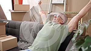 Middle age grey-haired man sitting on sofa stretching arms at new home