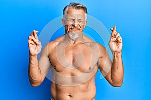 Middle age grey-haired man standing shirtless gesturing finger crossed smiling with hope and eyes closed