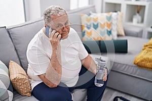 Middle age grey-haired man talking on smartphone holding bottle of water at home