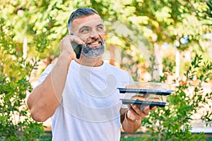 Middle age grey-haired man talking on the smartphone and holding take away food at the park